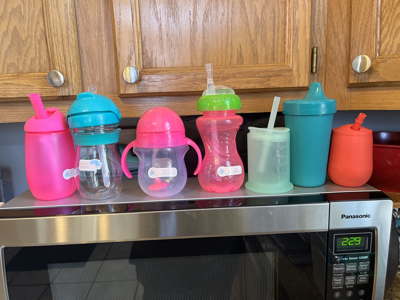 A row of colorful kids' sippy cups lined up on top of a microwave